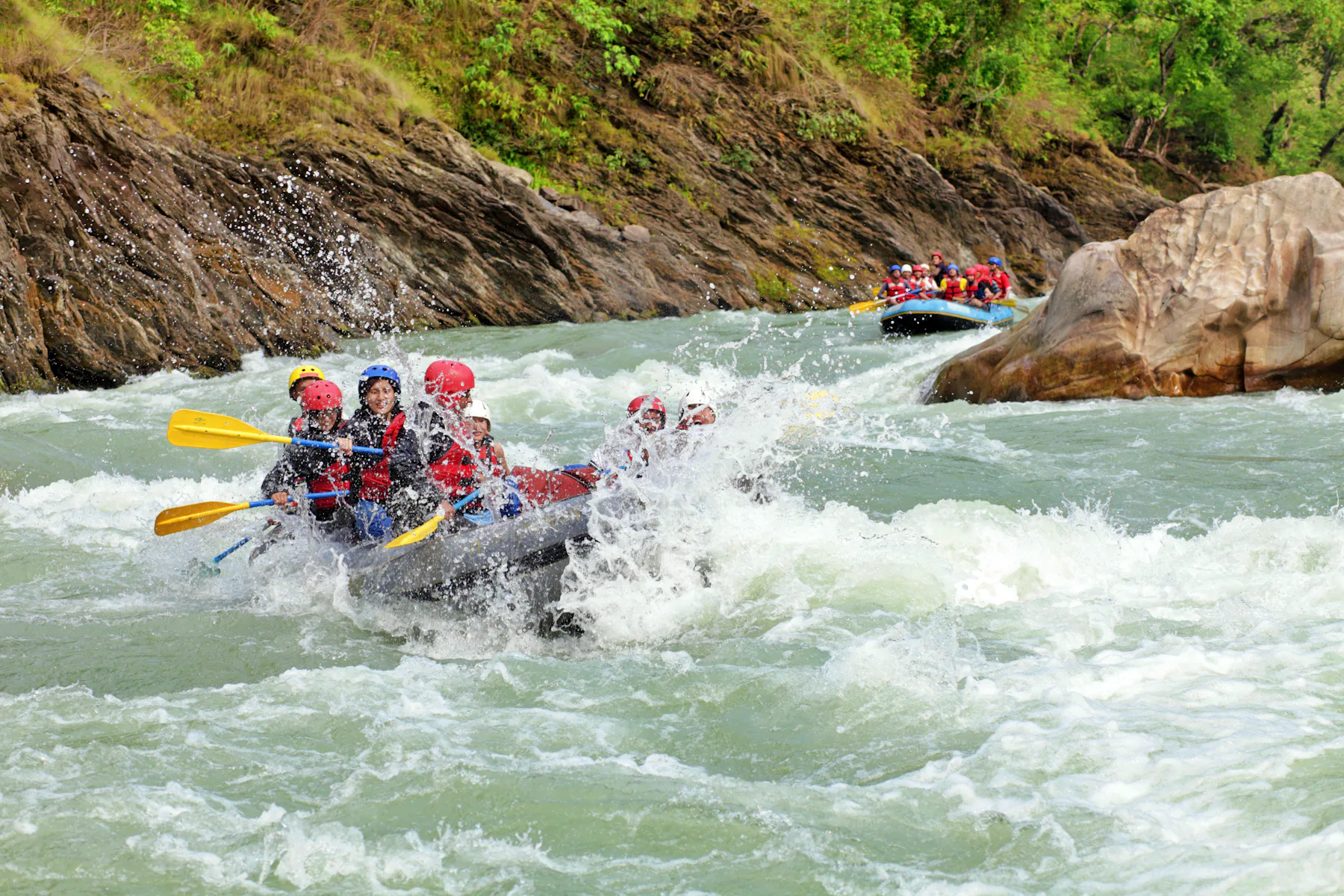 Raft crew in helmets paddling through white water below a rocky bank